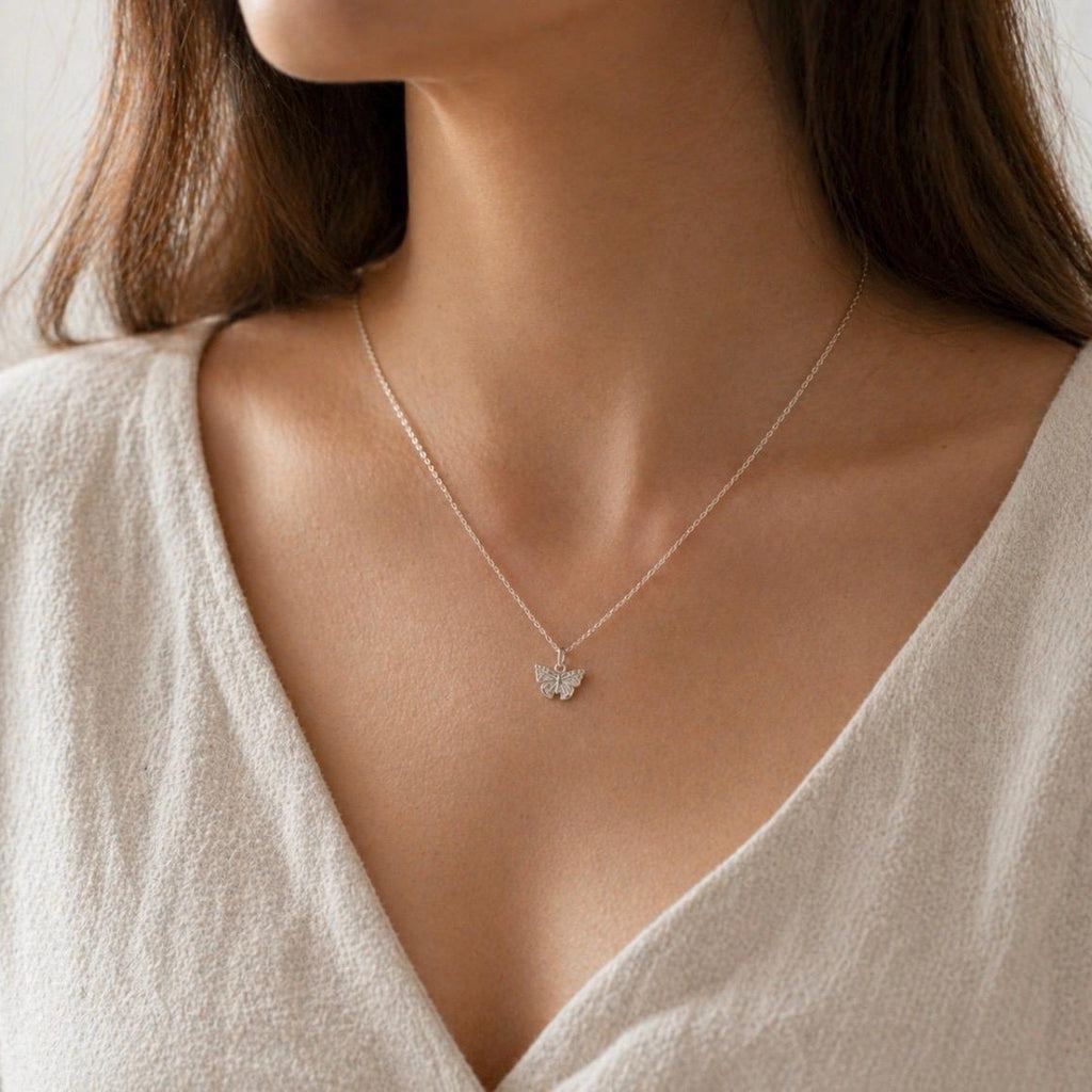 A delicate silver chain necklace with a small butterfly pendant, worn by a model in a close-up shot. The model is wearing a light beige V-neck top, and the background is a soft, neutral, light gray wall.