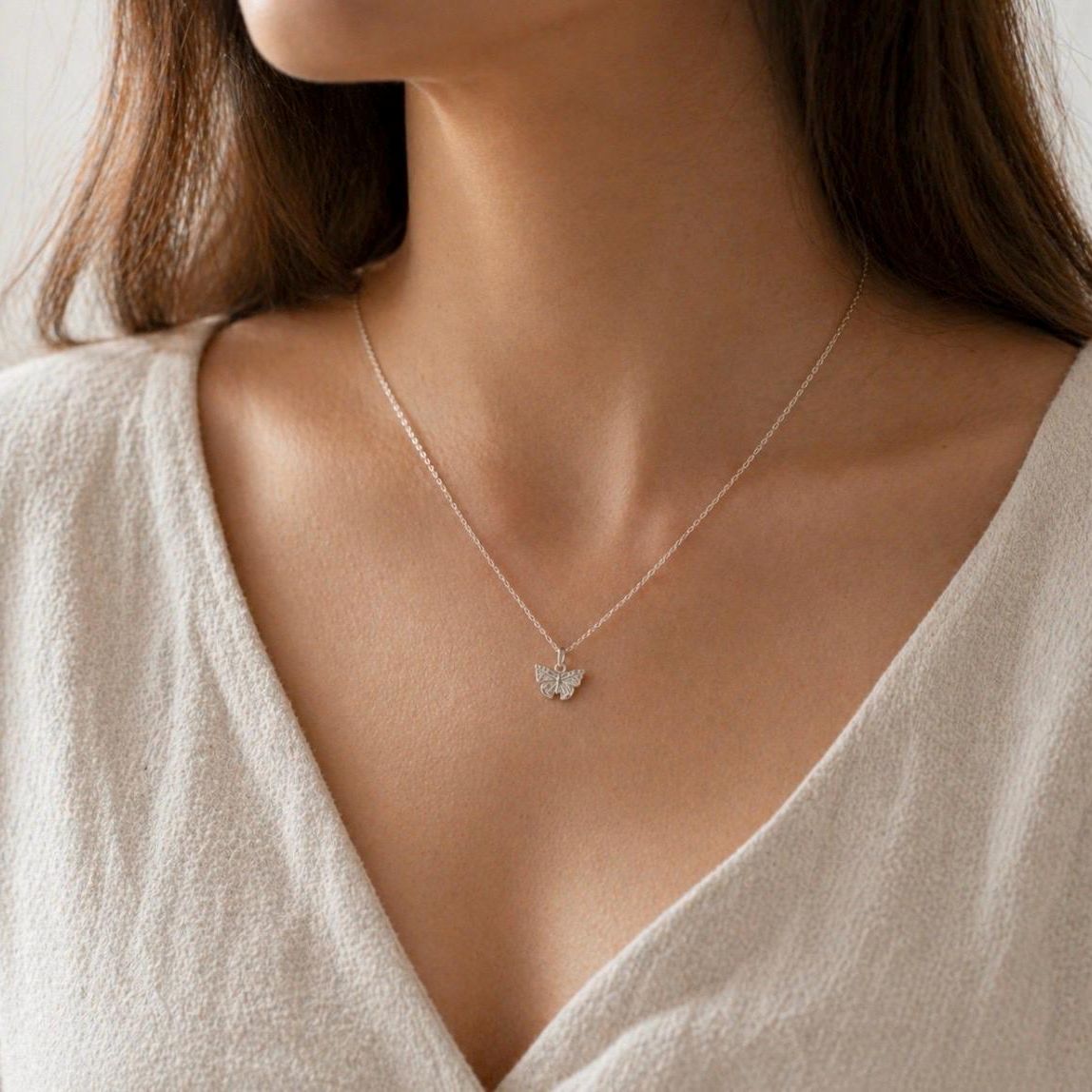 A close-up of a delicate silver chain necklace with a small butterfly pendant, worn by a model in a light beige V-neck top.