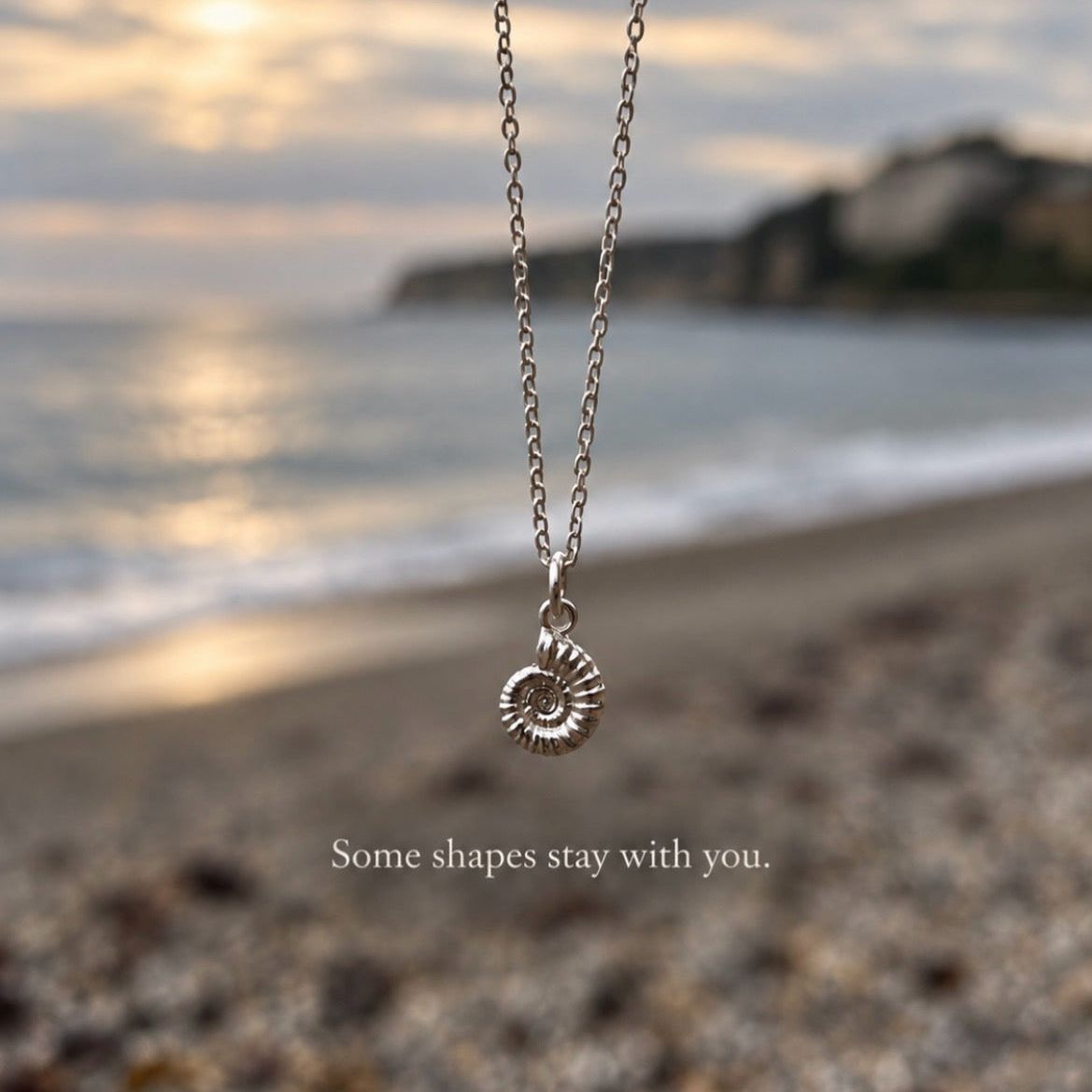 A silver chain necklace with a spiral ammonite pendant is shown hanging over a blurred beach shoreline at sunset. The text 'Some shapes stay with you.