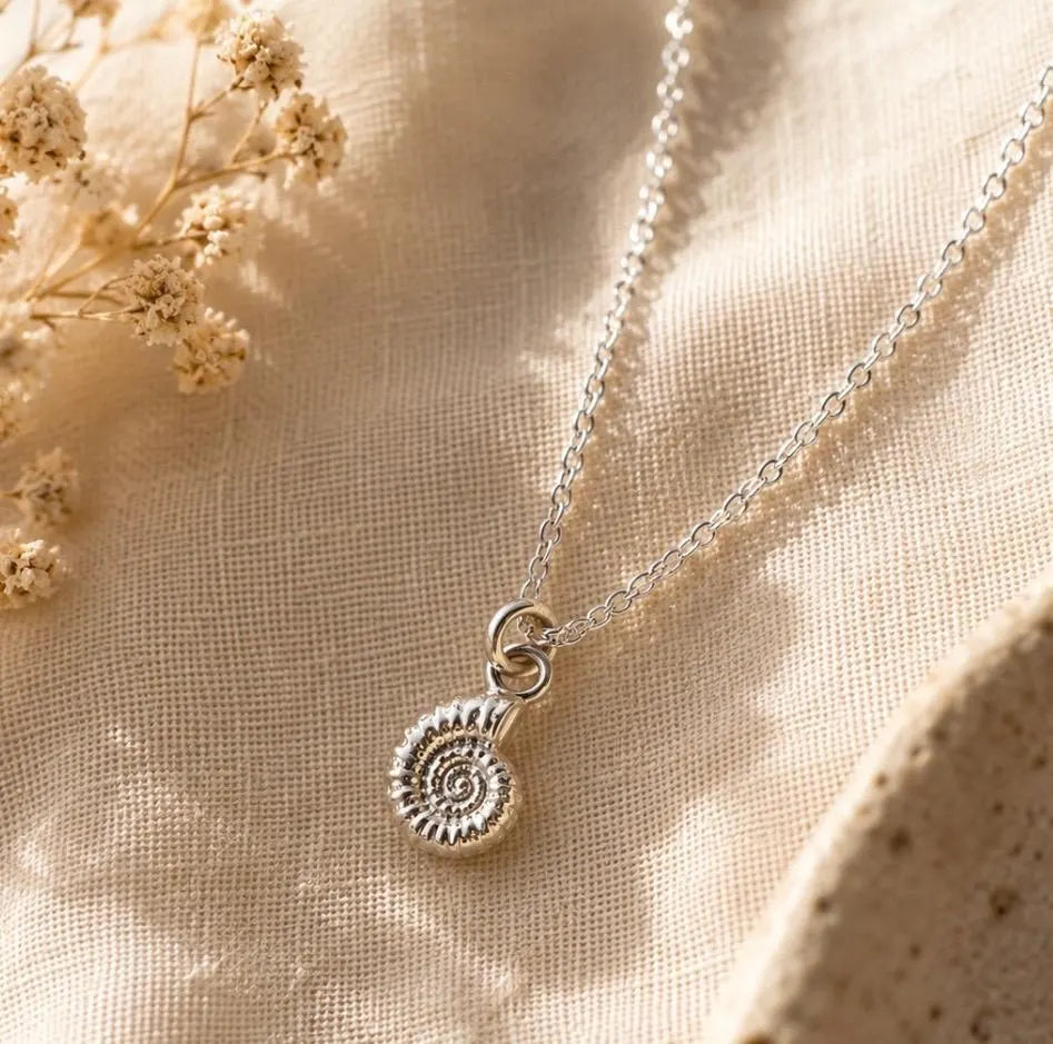 A silver chain necklace with a spiral ammonite pendant, styled on a textured beige linen fabric with dried flowers.