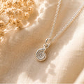 A silver chain necklace with a small spiral ammonite pendant, styled on a beige textured fabric with dried flowers in soft warm sunlight.
