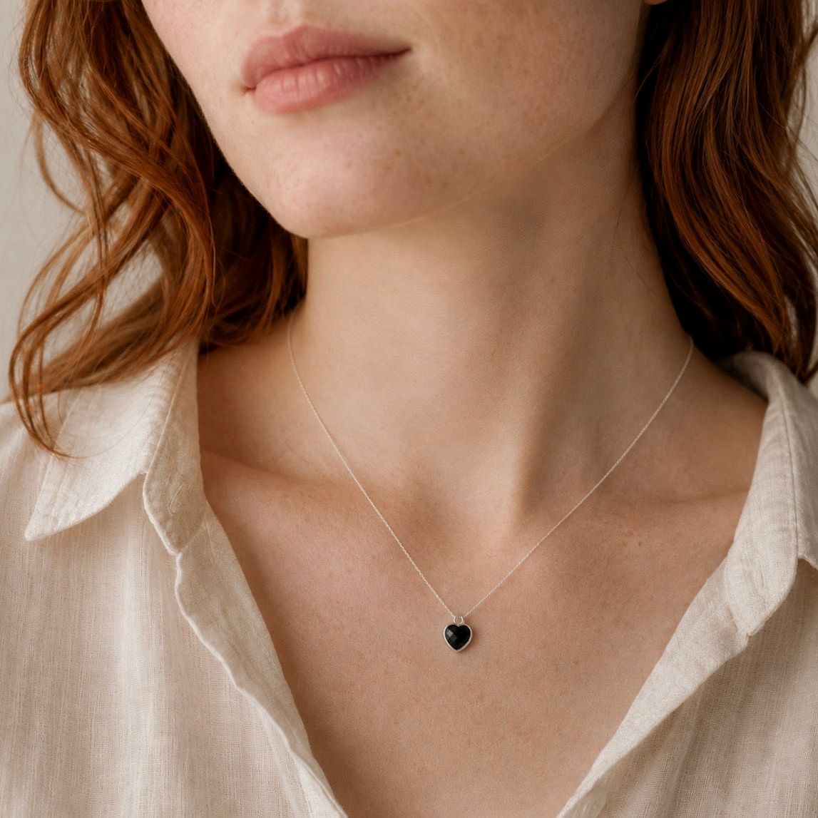 A close-up of a woman wearing a delicate silver chain necklace with a small black onyx heart-shaped pendant, paired with a light beige linen shirt.