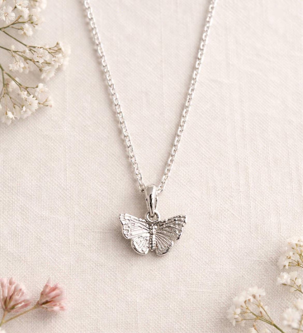 A silver butterfly pendant necklace on a delicate silver chain, displayed on a textured cream linen fabric with small white and pink baby's breath flowers.