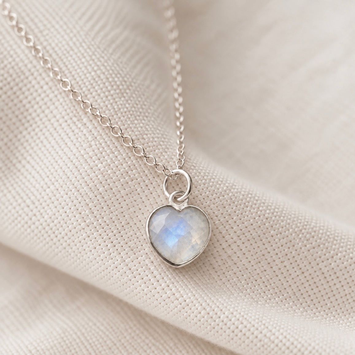 A silver heart pendant necklace with a milky white iridescent moonstone stone, resting on a textured beige fabric background.